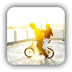 Two people riding together on a bicycle at sunset against a waterfront railing